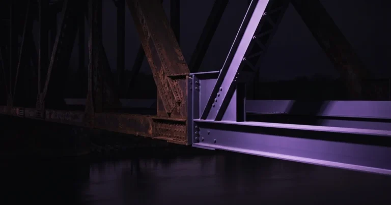 Estructura de acero oxidada de un puente, con vigas y remaches a la izquierda y vigas nuevas en tono púrpura a la derecha, sobre agua oscura de noche